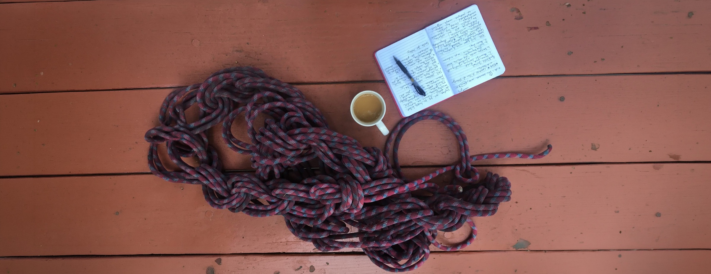 A coiled rope, an open journal, and a cup of coffee on a wooden deck