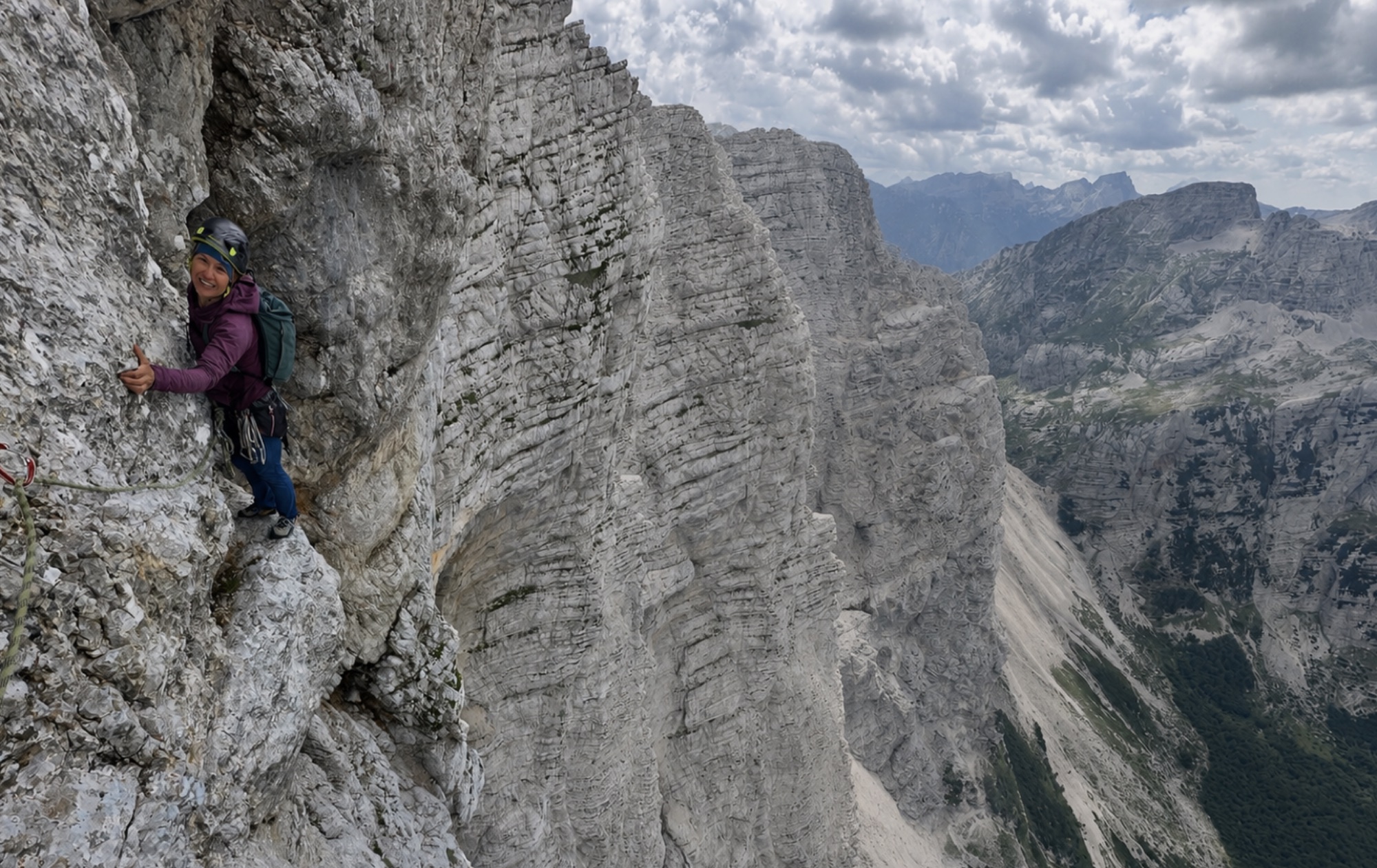 A climber smiling on a vast grey limestone wall high on the Triglav north face, a deep valley beyond