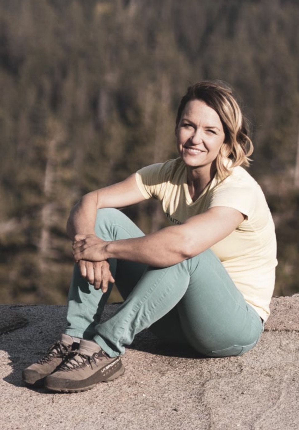 Marta sitting on rock in the sun, smiling, forest behind