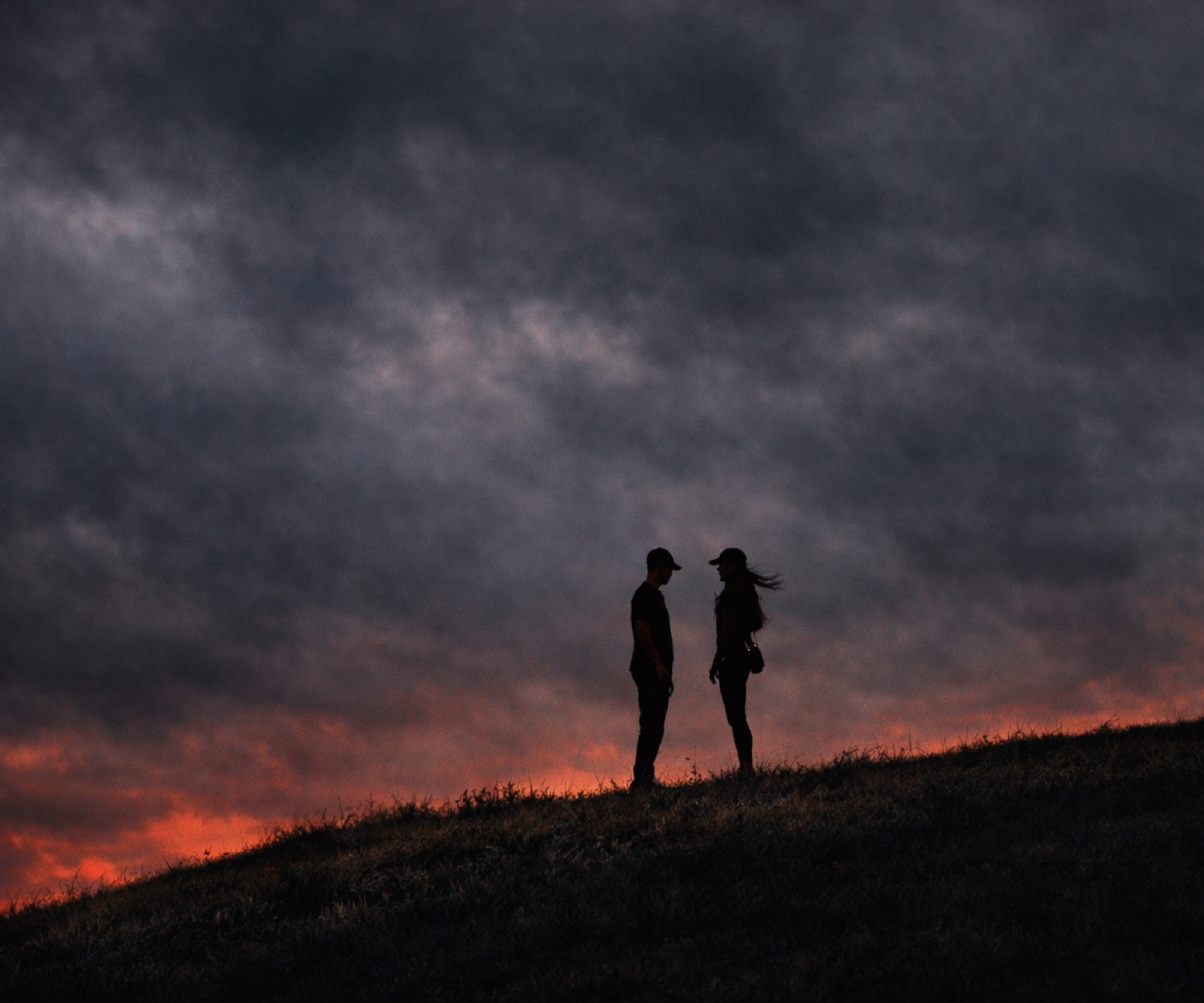 A couple facing each other in silhouette on a hilltop against a dark, ember-lit dusk sky